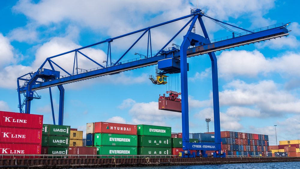 Blauwe portaalkraan van CTVrede - Steinweg die een container boven de kade van een containerterminal optilt, met gestapelde containers op de voorgrond